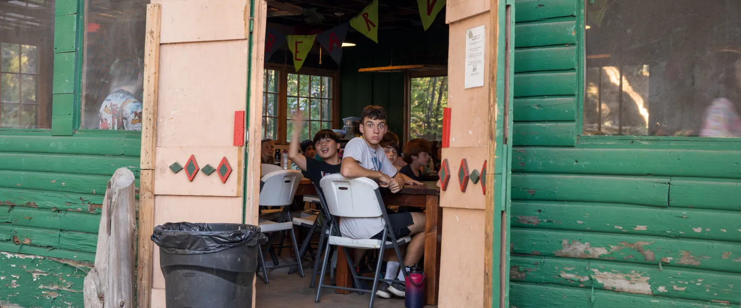 kids inside a mess hall, waving at the camera