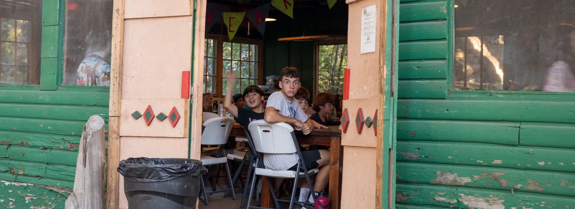 kids inside a mess hall, waving at the camera
